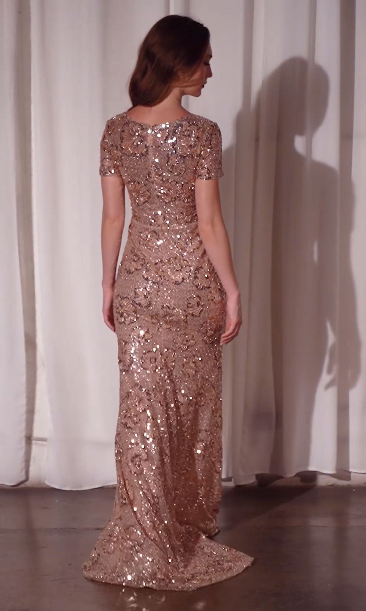A woman stands with her back to the camera, wearing the fitted, floor-length Ladivine CC3700 Rose Gold Prom Dress adorned with floral sequins and short sleeves. Her hair is down, and she’s in front of white curtains on a concrete floor.